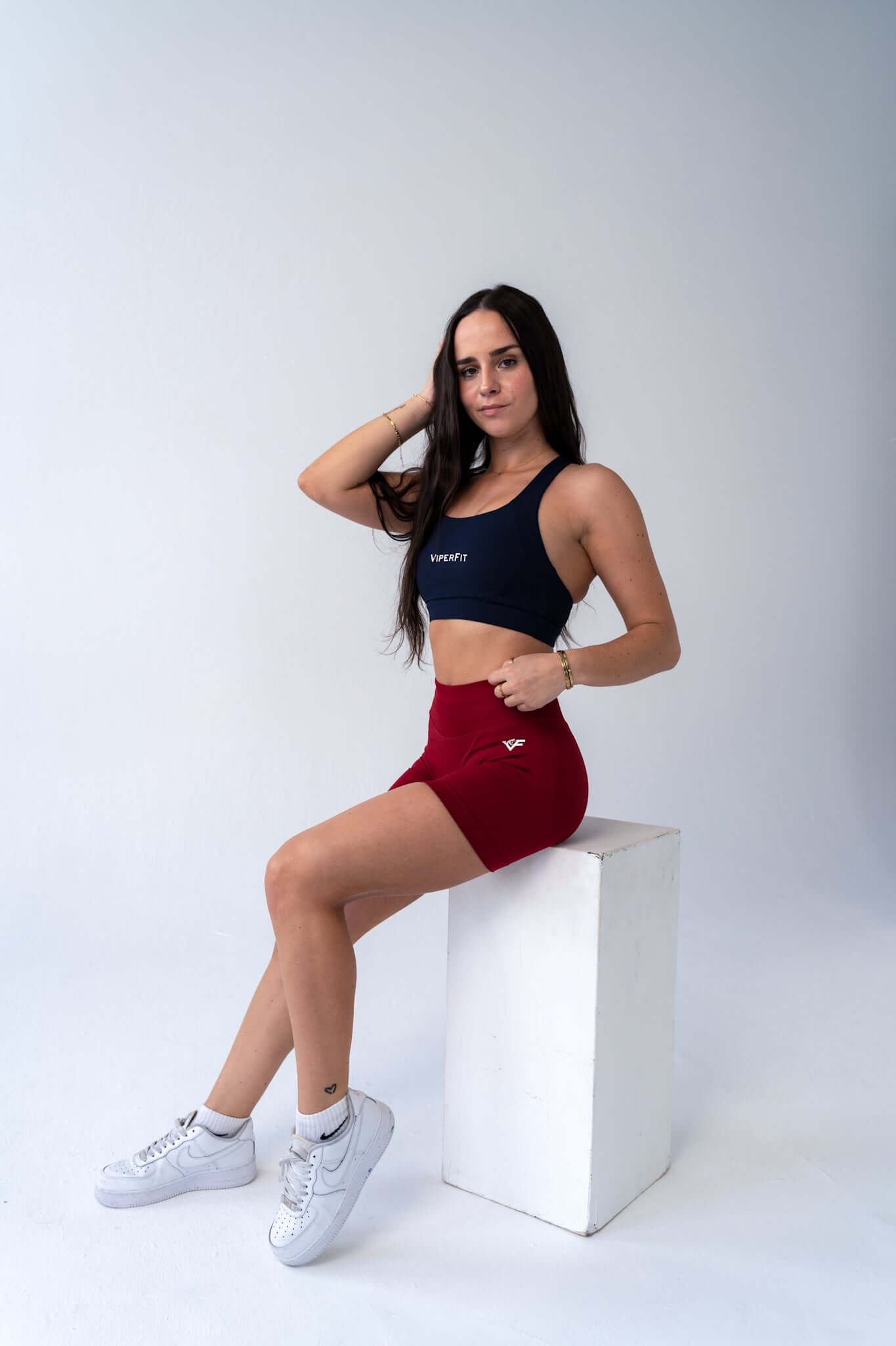Model seated in ViperFit Luxe Pro Shorts - Wine with a navy blue pulse sports bra and white trainers.